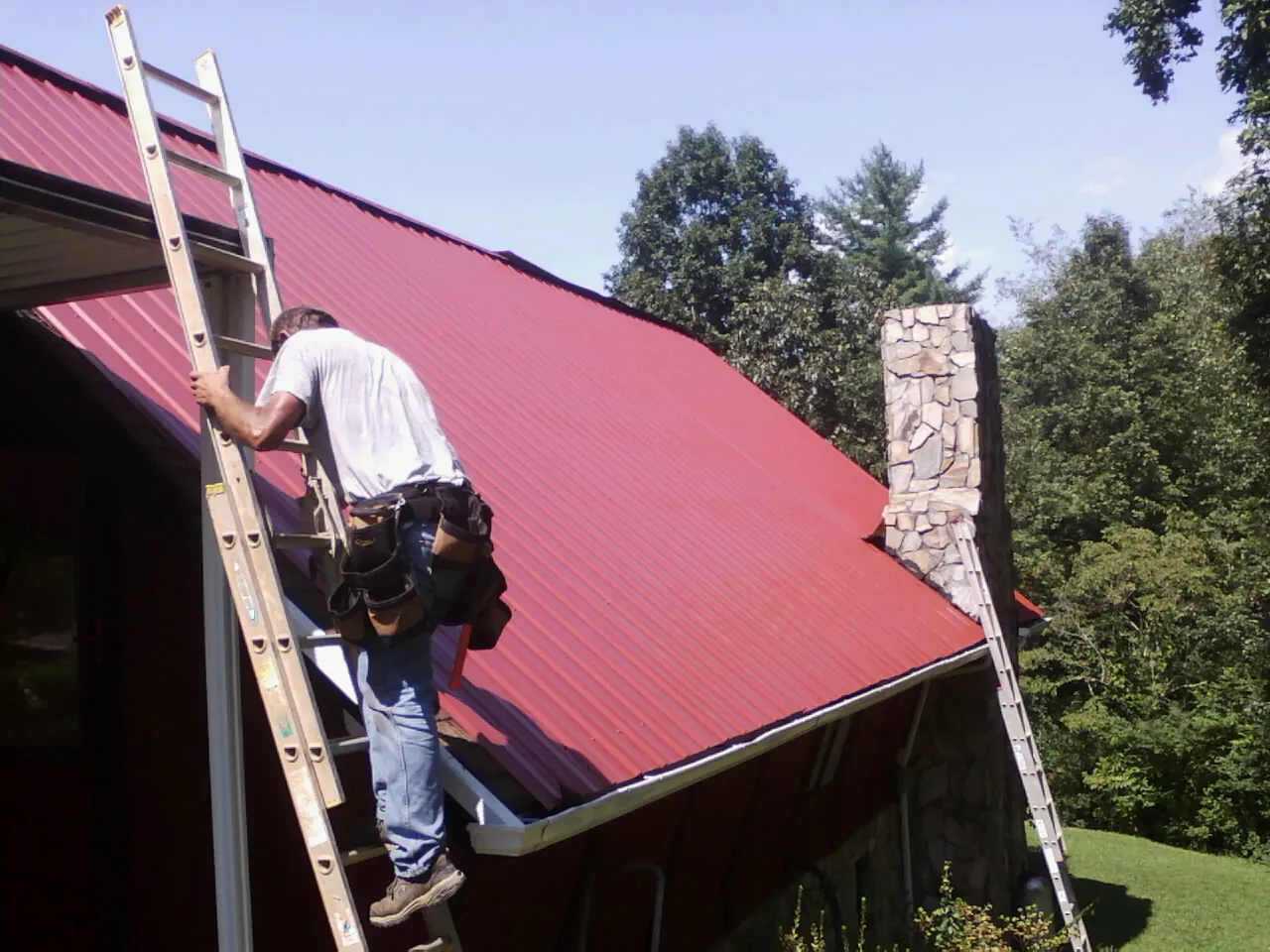 Roofer on a ladder beside a red metal roof and stone chimney — Soffit & Fascia Repair by Roofing Brothers in Hillsdale